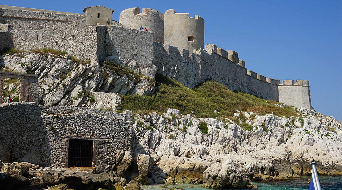 A stone castle sitting atop a white rocky island on blue water with fortress-like walls and numerous towers